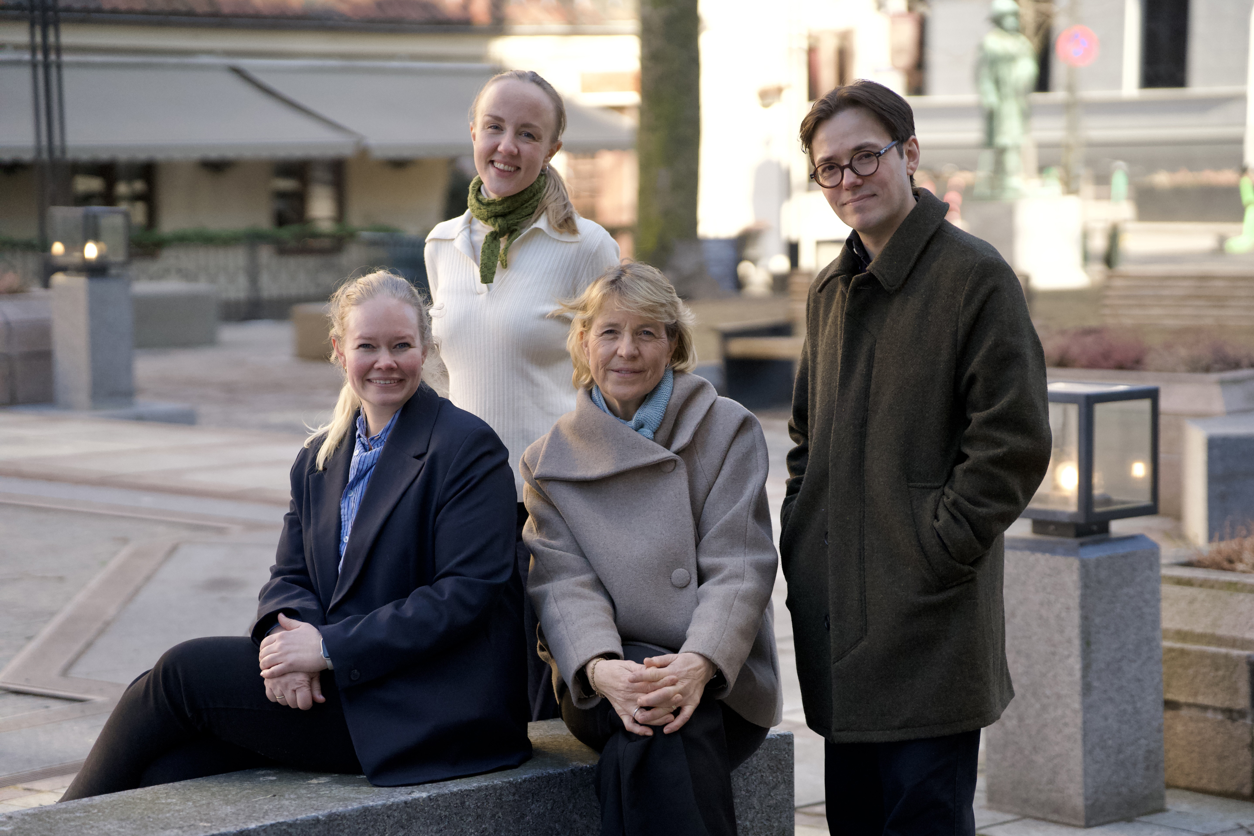 Fire personer med yttertøy står ute i et byrom og ser mot kameraet med avslappet og profesjonelt ansiktsuttrykk.