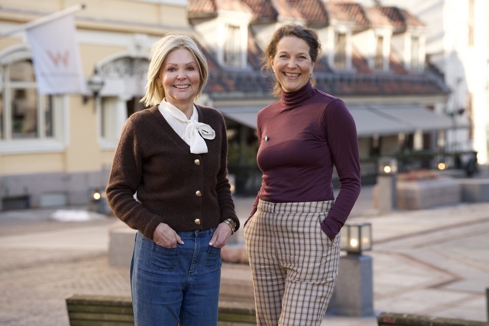 To kvinner står avslappet med hendene i lommen og smiler til kameraet. Bakgrunnen antyder en bygate eller et moderne bylandskap.